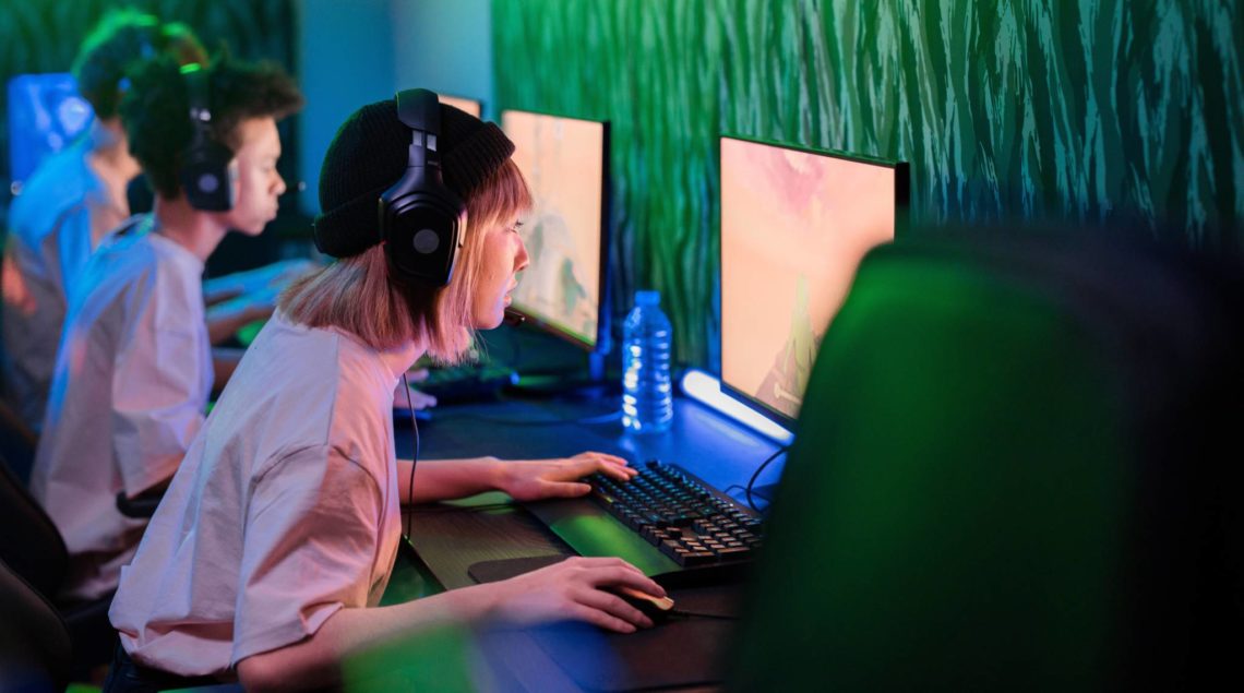 A woman with a headset is leaning towards her PC which shows a video game on the screen. She is in a room with two men who are also staring at their screens. The room is dimly lit with fluro green lights.
