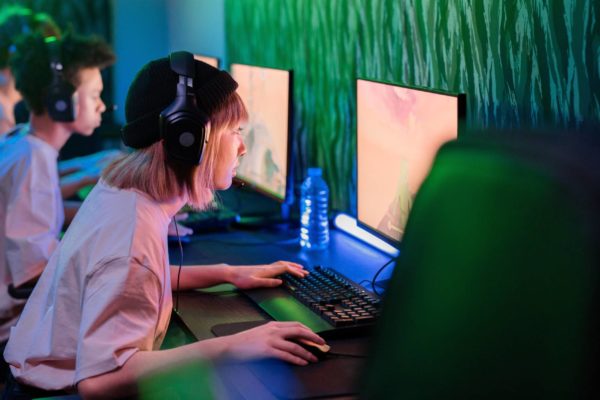 A woman with a headset is leaning towards her PC which shows a video game on the screen. She is in a room with two men who are also staring at their screens. The room is dimly lit with fluro green lights.