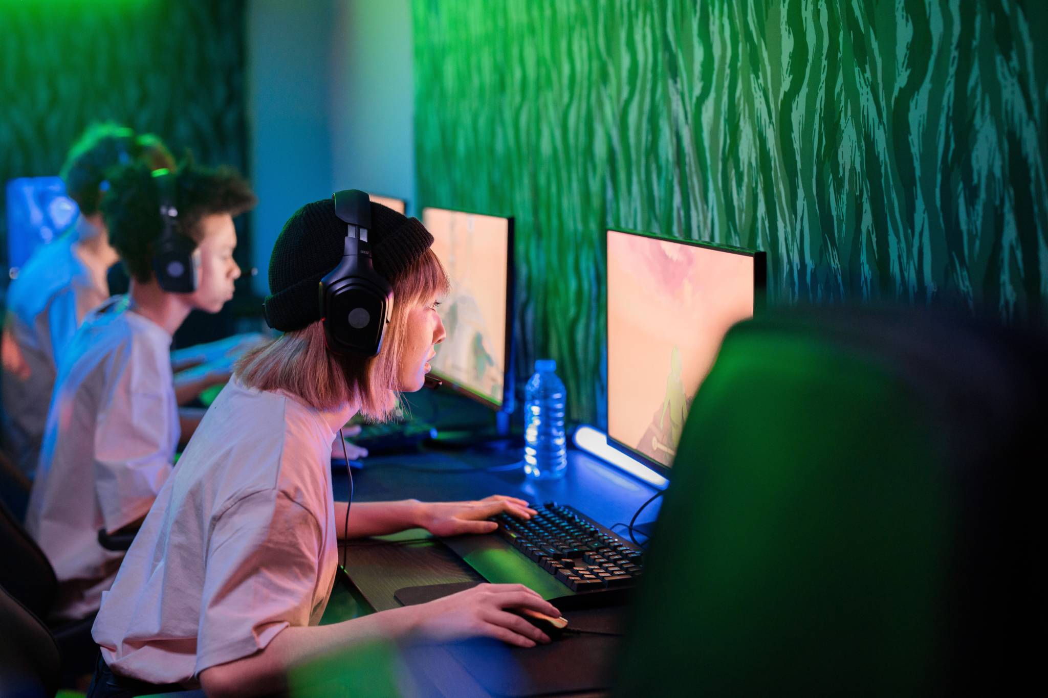 A woman with a headset is leaning towards her PC which shows a video game on the screen. She is in a room with two men who are also staring at their screens. The room is dimly lit with floor green lights.