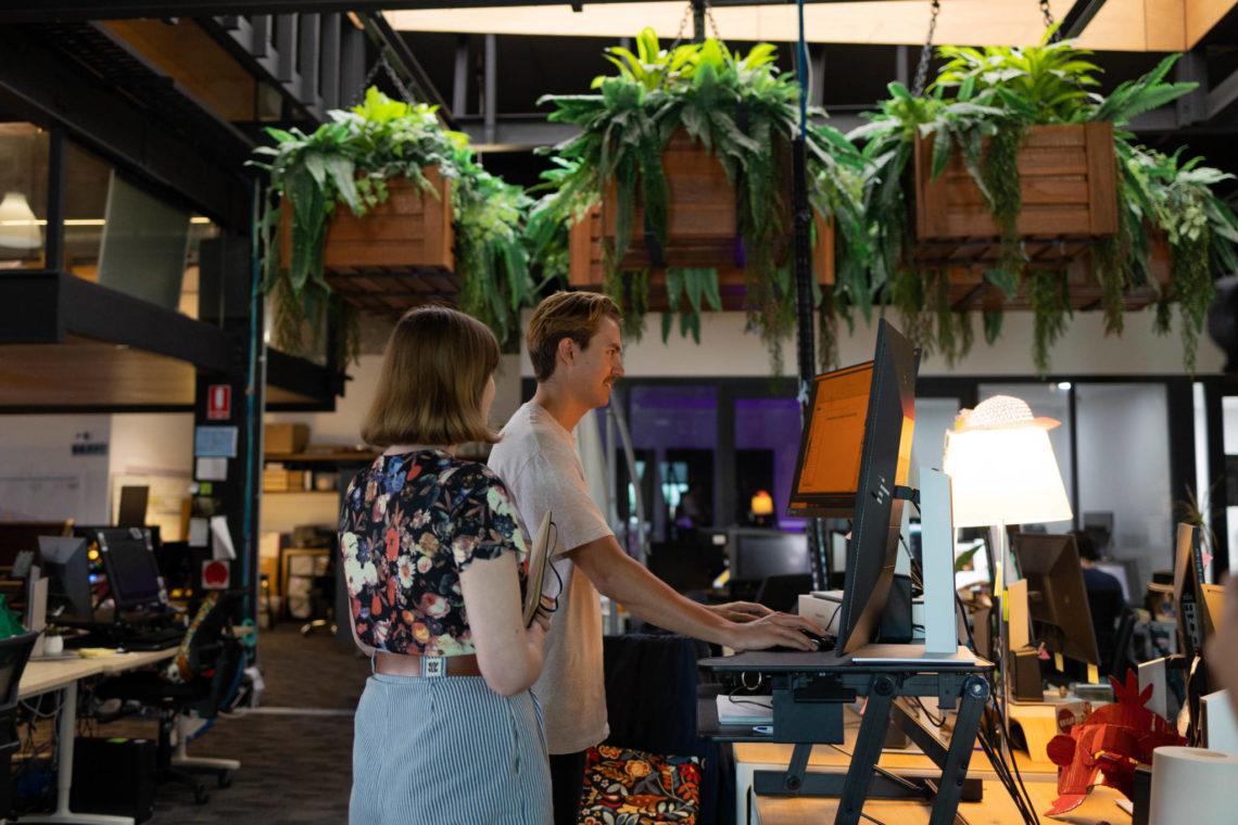 A woman and man are standing at a stand-up desk iand looking at a computer in a leafy, industrial-looking office