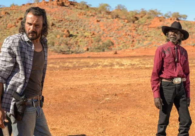 Two men stand outside. The earth is red and the image is from film MYSTERY ROAD