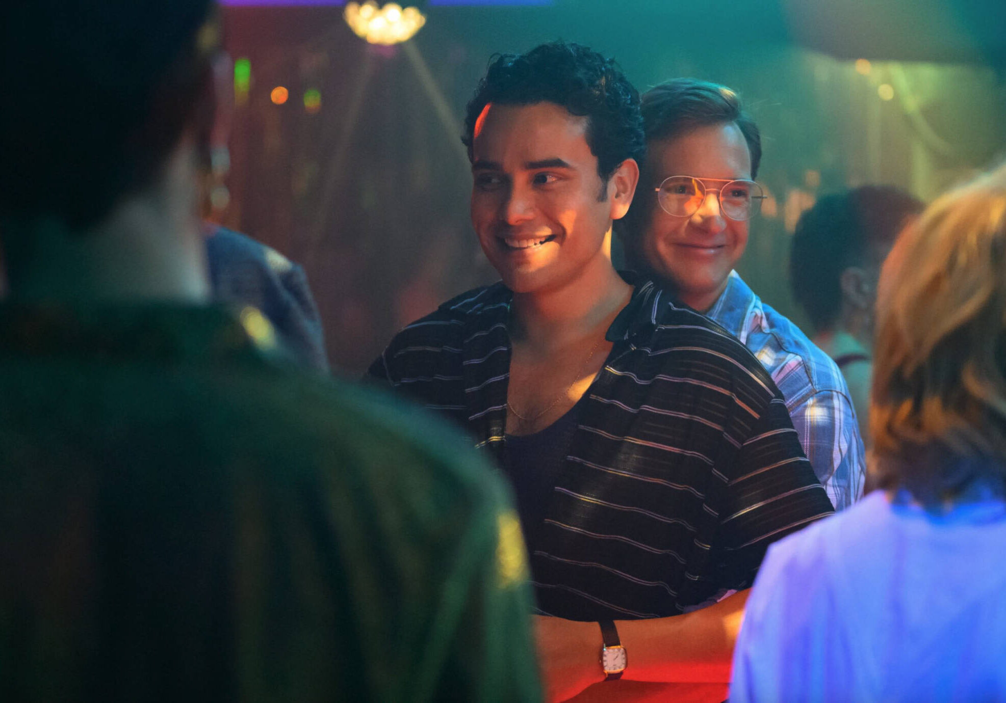 A photo of two men in a club talking to other people. One of the men is hugging the other from behind.