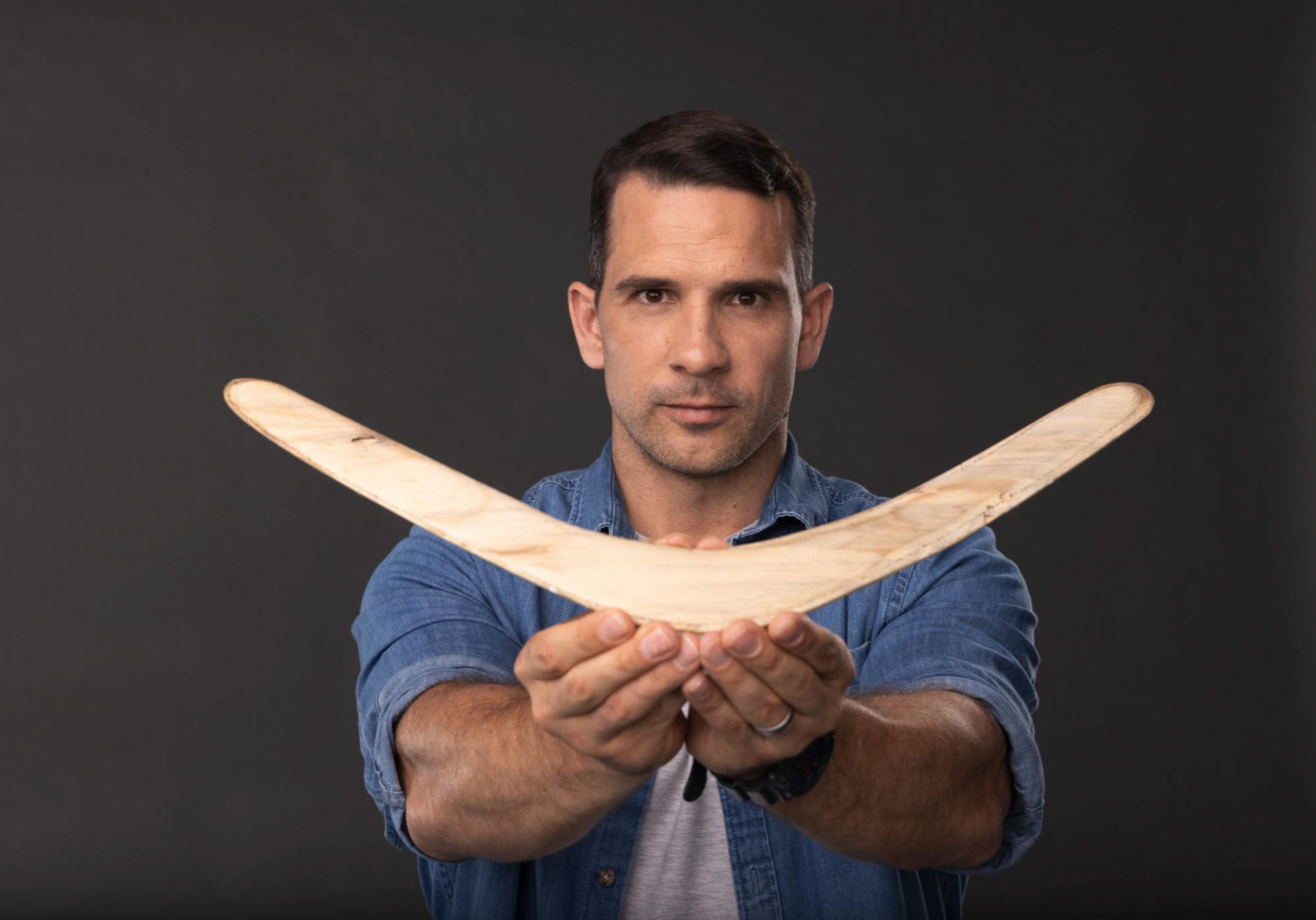 Phillip Breslin looks at the camera and holds a boomerang as part of FIRST WEAPONS