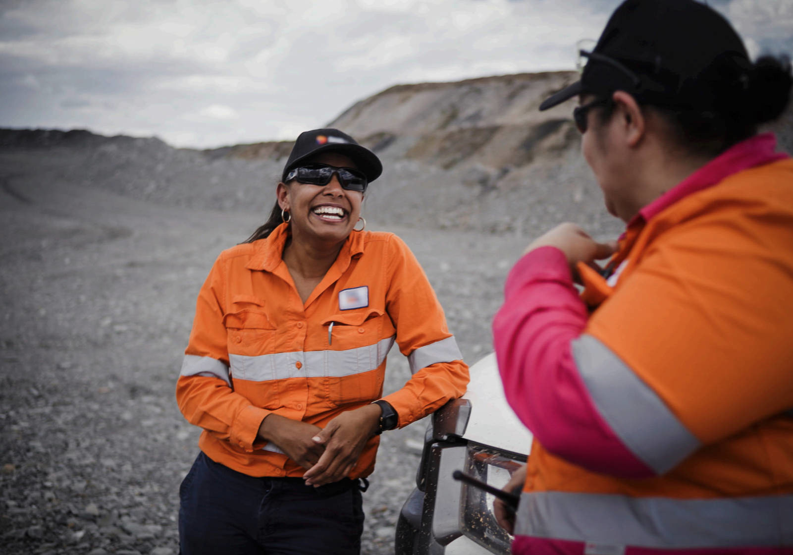 A woman is laughing and smiling at another woman. They are both in high vis and standing outside. This is from the documentary SISTAS IN MINING