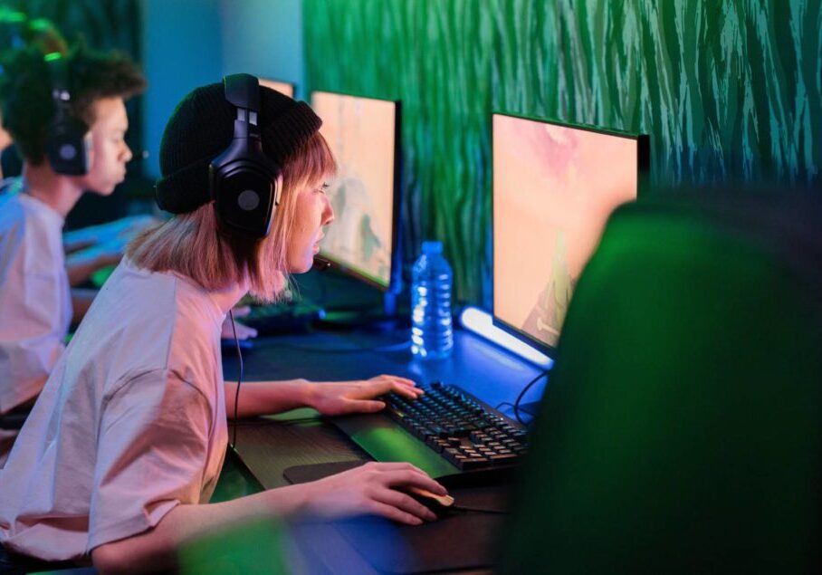 A woman with a headset is leaning towards her PC which shows a video game on the screen. She is in a room with two men who are also staring at their screens. The room is dimly lit with fluro green lights.