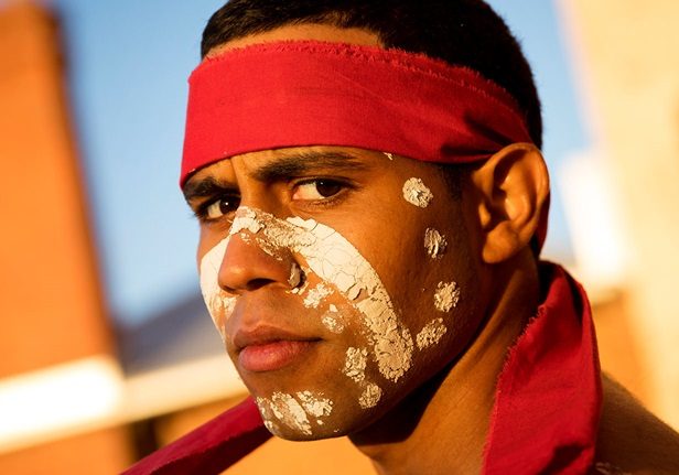 A man with white paint on his face and wearing a red bandana looks at the camera from series WRONG KIND OF BLACK